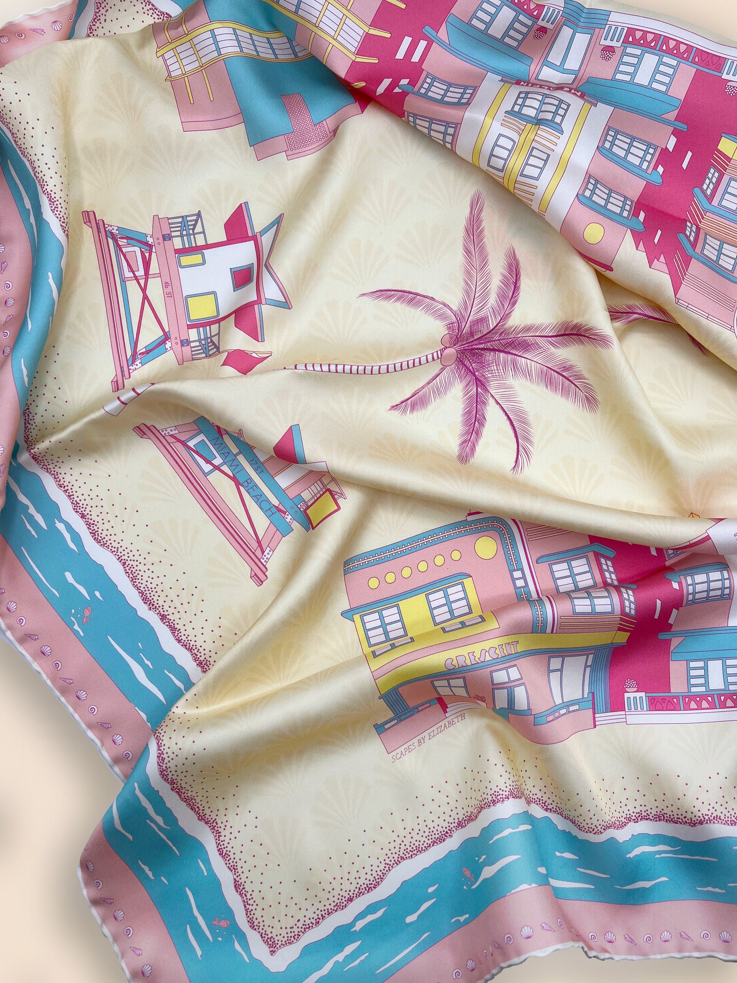 Close-up of the Rosy Splash Miami Beach silk scarf showcasing hand-drawn Art Deco architecture, iconic miami beach lifeguard towers, palm trees, a subtle shell pattern, and a decorative seashell border.