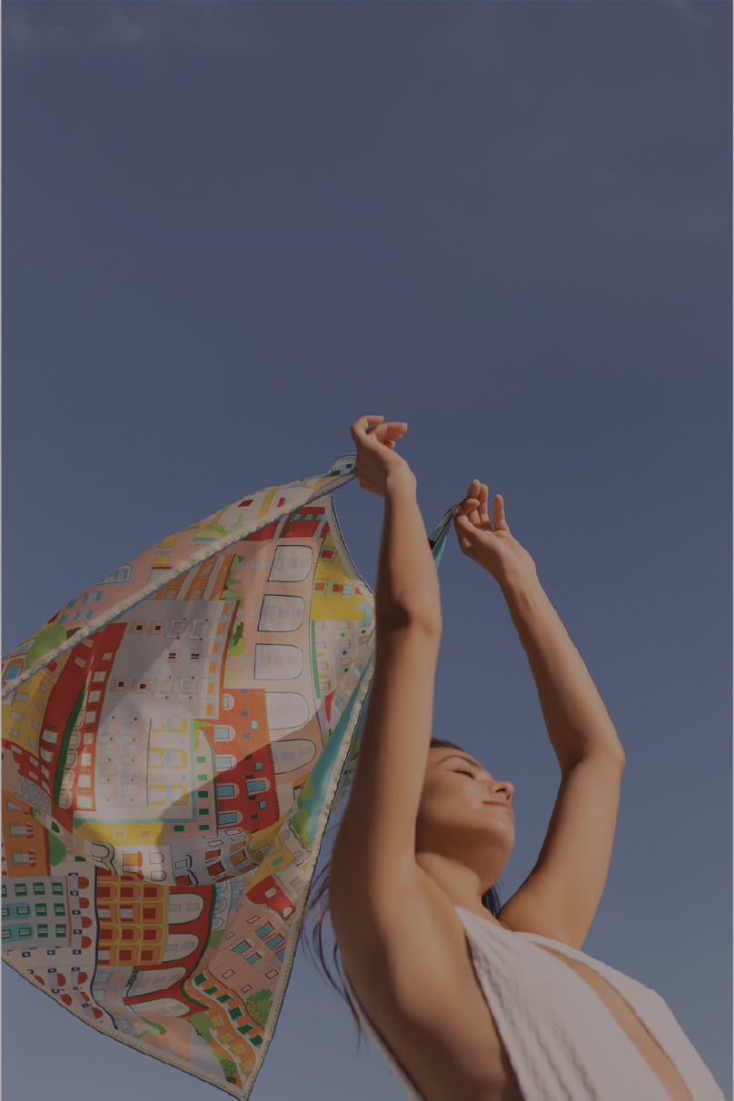 A woman outdoors against a clear blue sky holds up a colorful patterned amali coast inspired scarf or shawl with both hands. She is wearing a light sleeveless top.