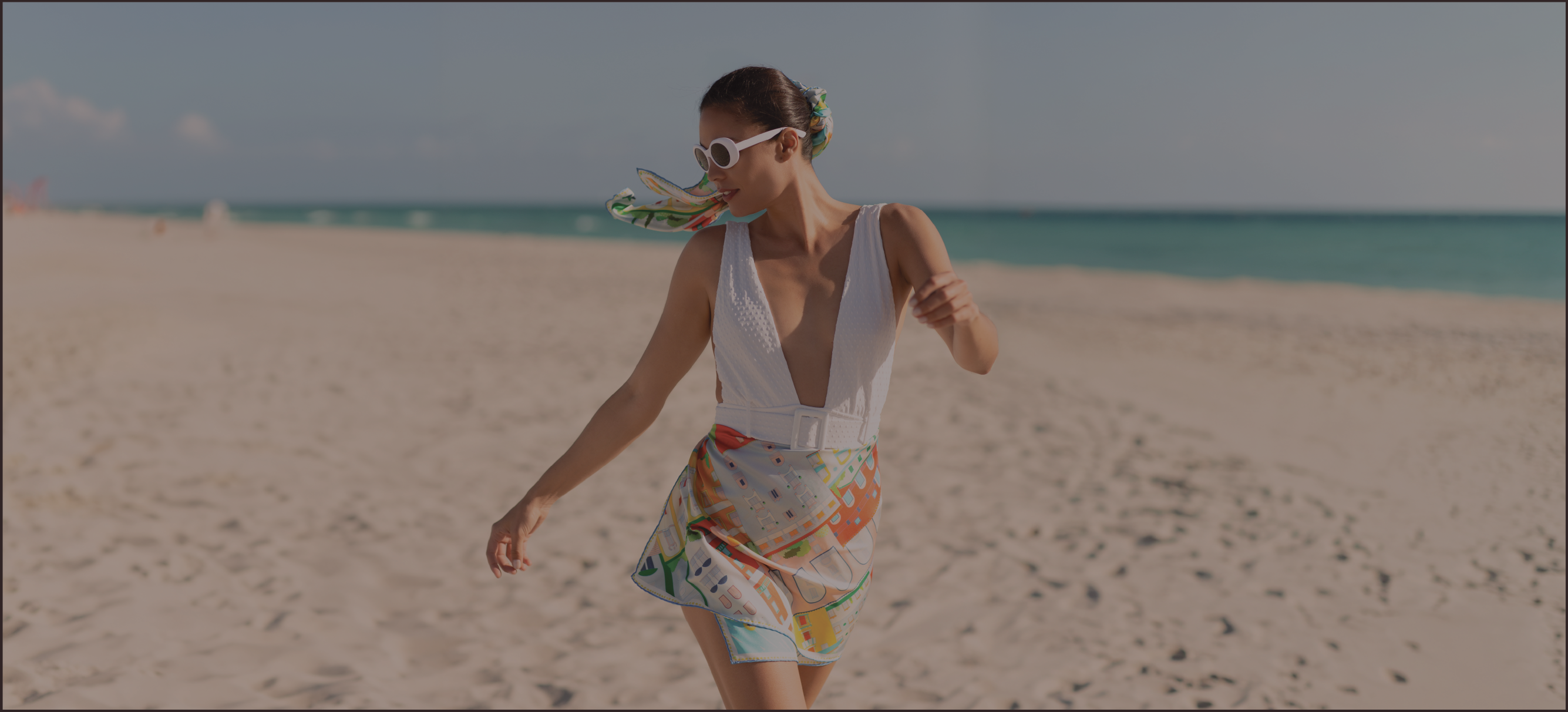 A woman walking on a sandy beach wearing a white swimsuit with a colorful amalfi coast inspired silk scarf and white sunglasses.