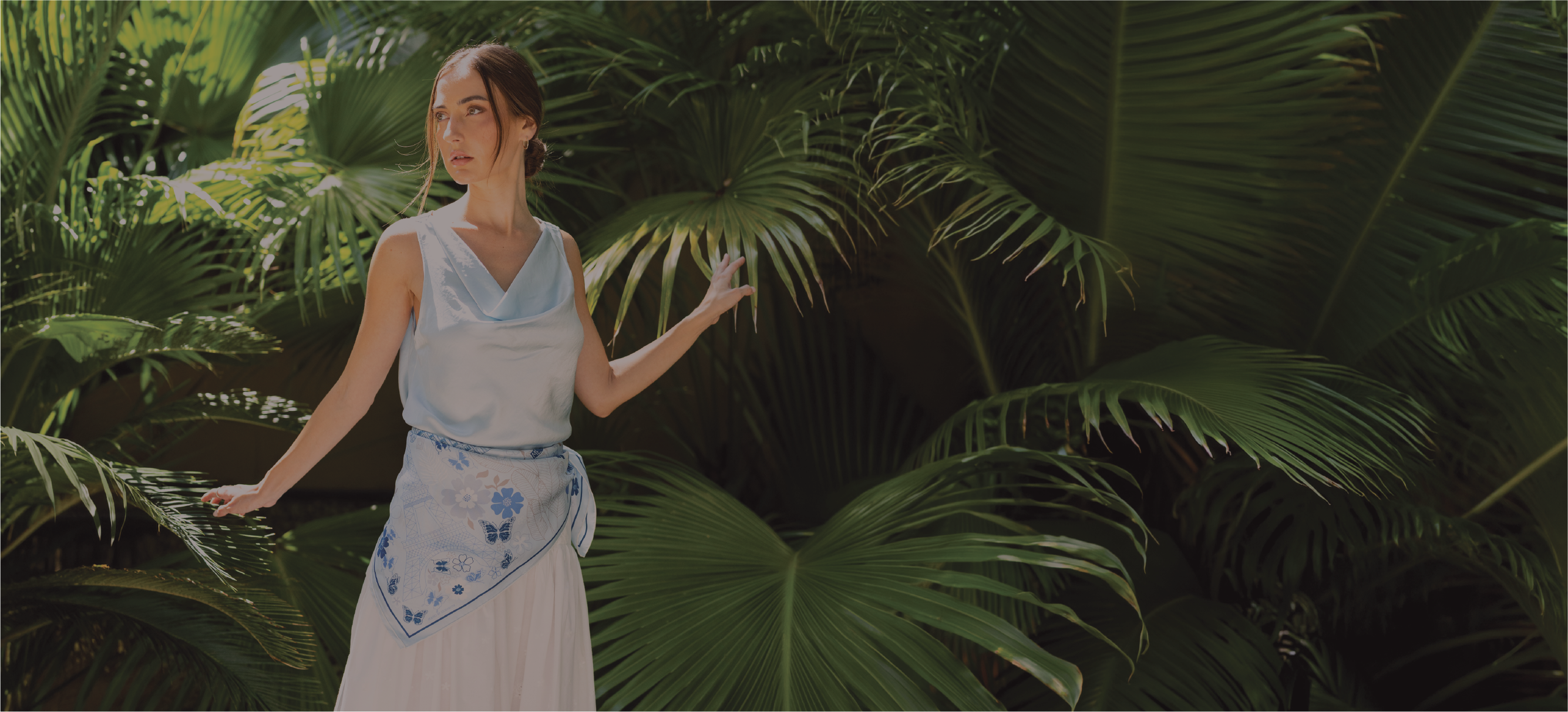 A woman wearing a light blue sleeveless top and a white skirt with a blue and white paris inspired silk scarf stands among large green tropical palm leaves in a shaded outdoor setting.