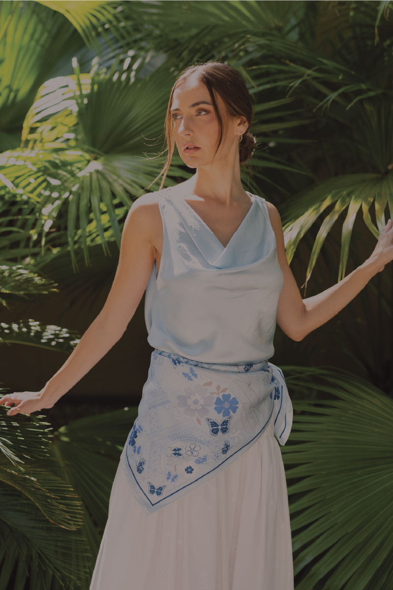 A woman wearing a light blue sleeveless top and a white skirt with a blue and white paris inspired silk scarf tied around her waist, posing outdoors in front of lush green palm leaves.