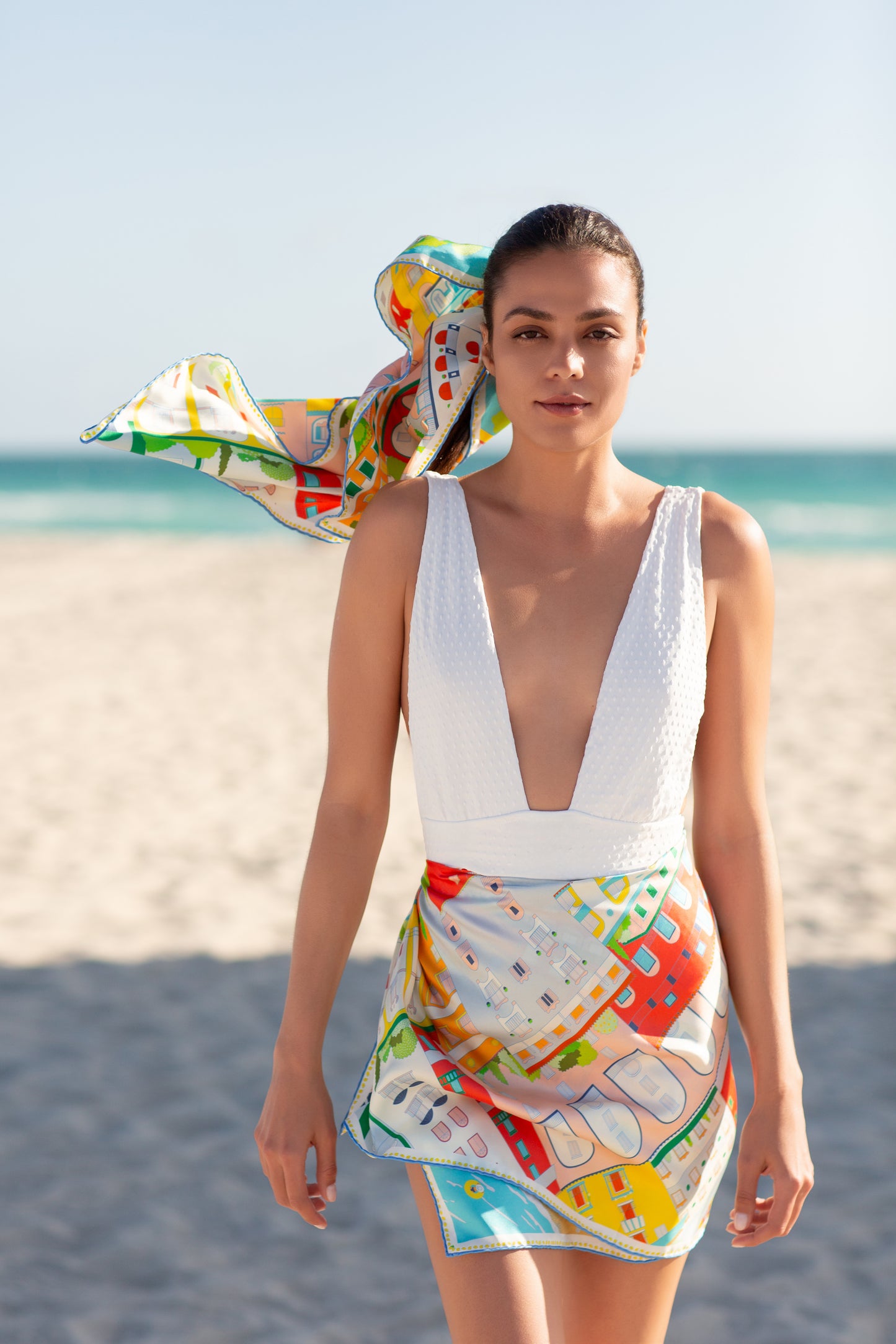 Woman showcasing the versatility of the Amalfi Coast silk scarf, styled as a beach cover-up skirt and hair accessory, featuring hand-drawn illustrations of Positano, colorful cliffside buildings, and Mediterranean coastal scenery.