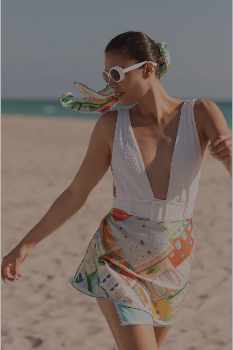 A woman walking on a sandy beach wearing a white swimsuit with a colorful amalfi coast inspired silk scarf wrapped as a skirt and white sunglasses.