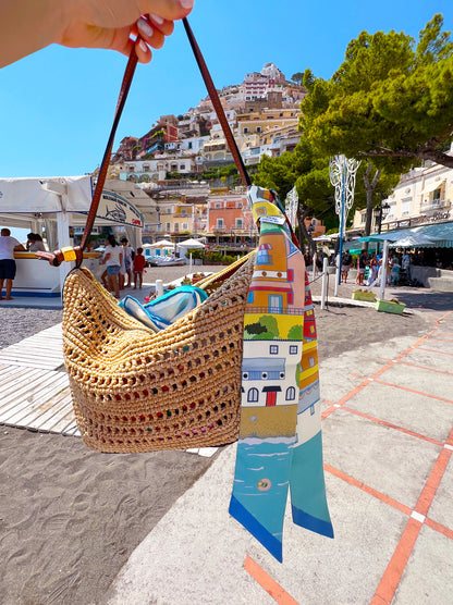 Citrus Splash Amalfi Coast silk twilly featuring hand-drawn illustrations inspired by Positano and colorful Mediterranean villages tied on a raffia handbag with Italy's Amalfi Coast in the background.