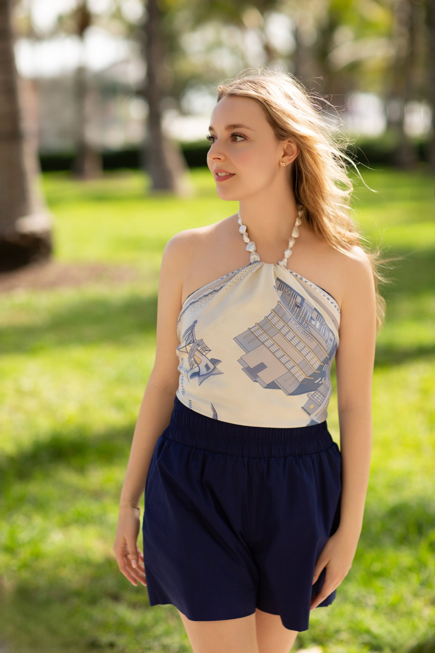 Woman wearing the Cool Blue Miami Beach silk scarf as a halter top featuring hand-drawn Art Deco architecture, lifeguard towers, palm trees, and coastal-inspired artwork in shades of blue.