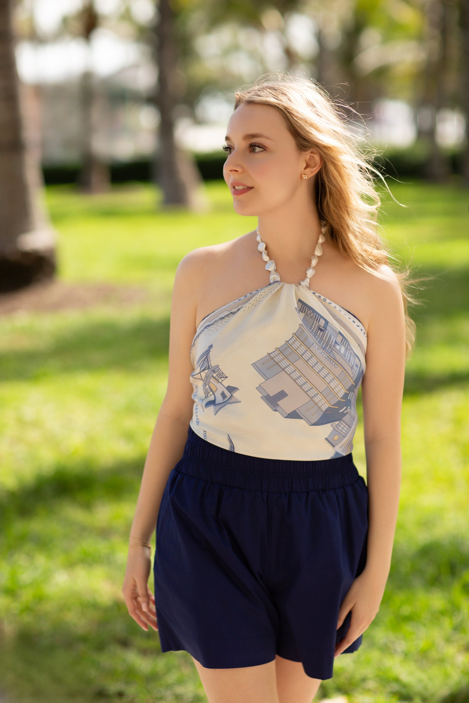 Woman wearing the Cool Blue Miami Beach silk scarf as a halter top featuring hand-drawn Art Deco architecture, lifeguard towers, palm trees, and coastal-inspired artwork in shades of blue.