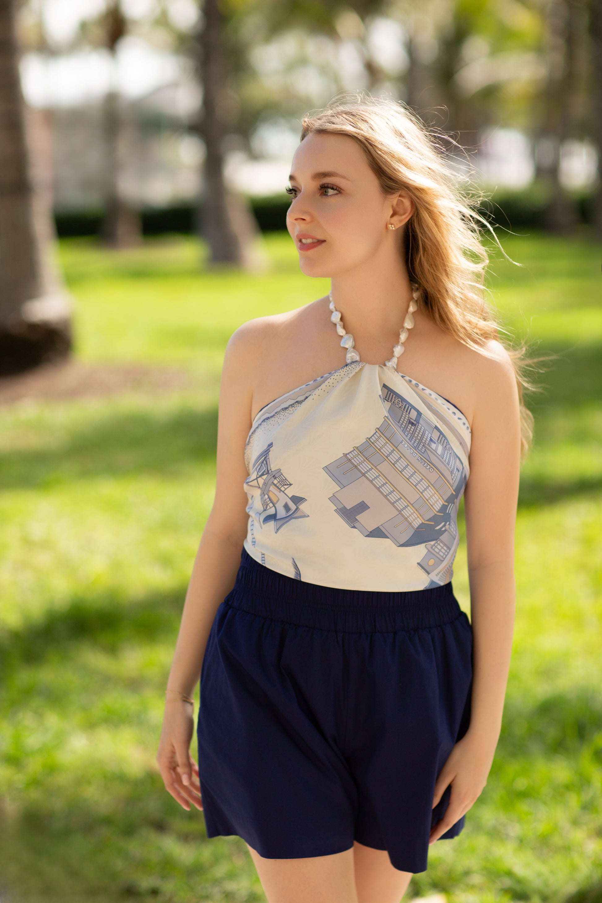 Woman wearing the Cool Blue Miami Beach silk scarf as a halter top featuring hand-drawn Art Deco architecture, lifeguard towers, palm trees, and coastal-inspired artwork in shades of blue.