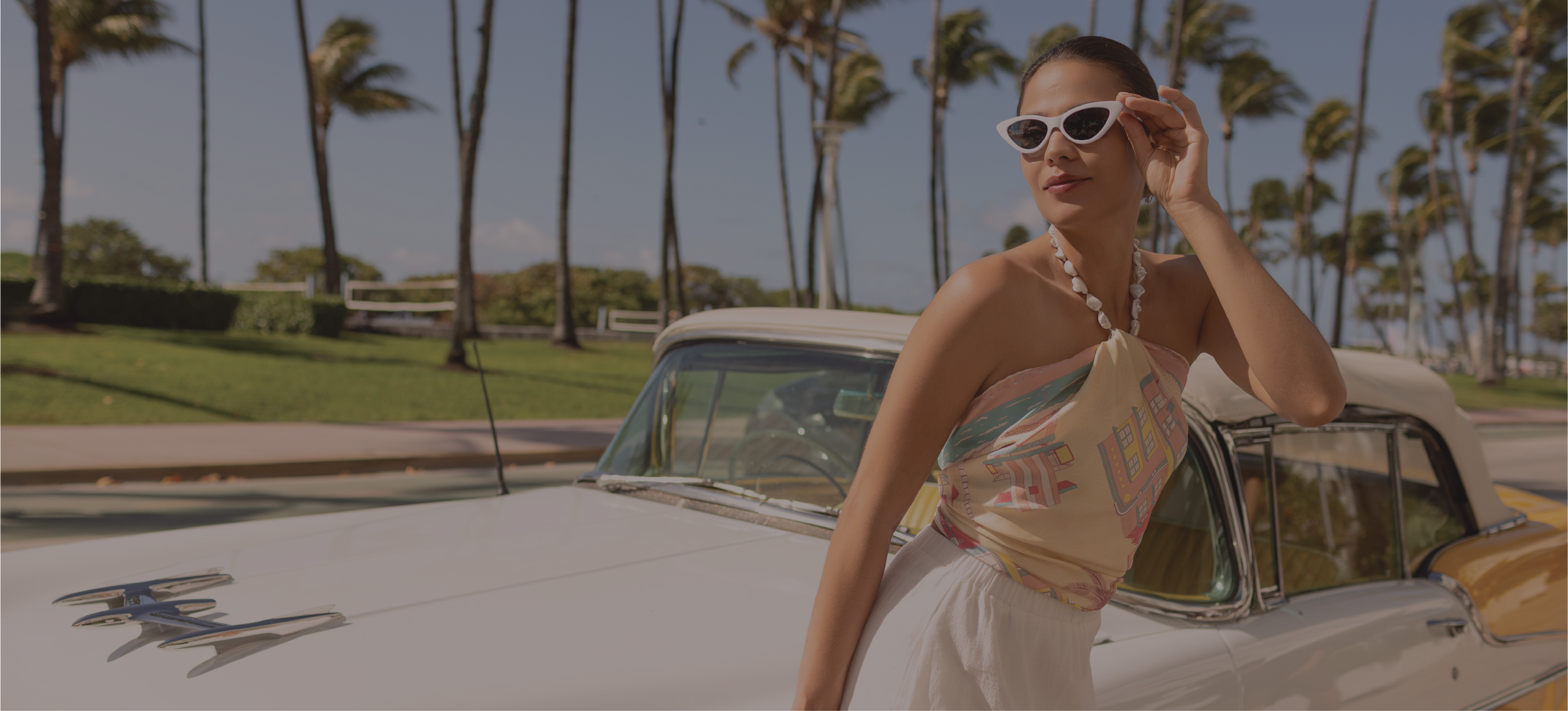 A woman wearing white sunglasses and a miami beach inspired silk scarf as a top leans against a white vintage car outdoors, with palm trees and a sunny sky in the background.