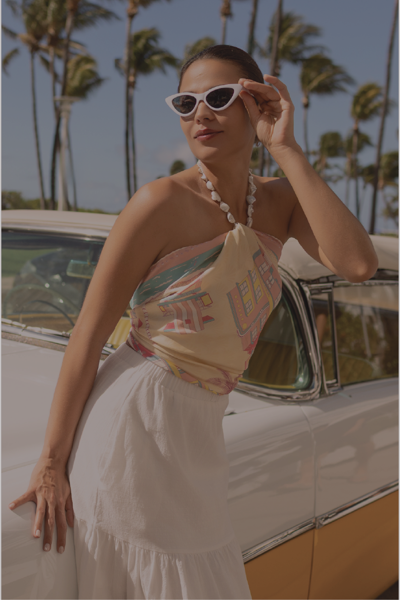 A woman wearing a miami beach inspired silk scarf tied as a halter top and a white skirt, posing next to a vintage white car with palm trees in the background.