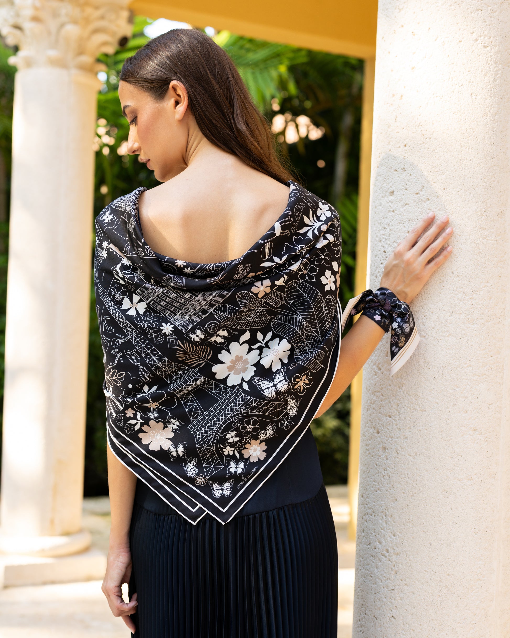 Woman wearing a black and white Paris silk scarf featuring hand-drawn Paris landmarks including the Eiffel Tower, Moulin Rouge, Arc de Triomphe, luxury boutiques, and fashion-inspired details styled as a shawl.