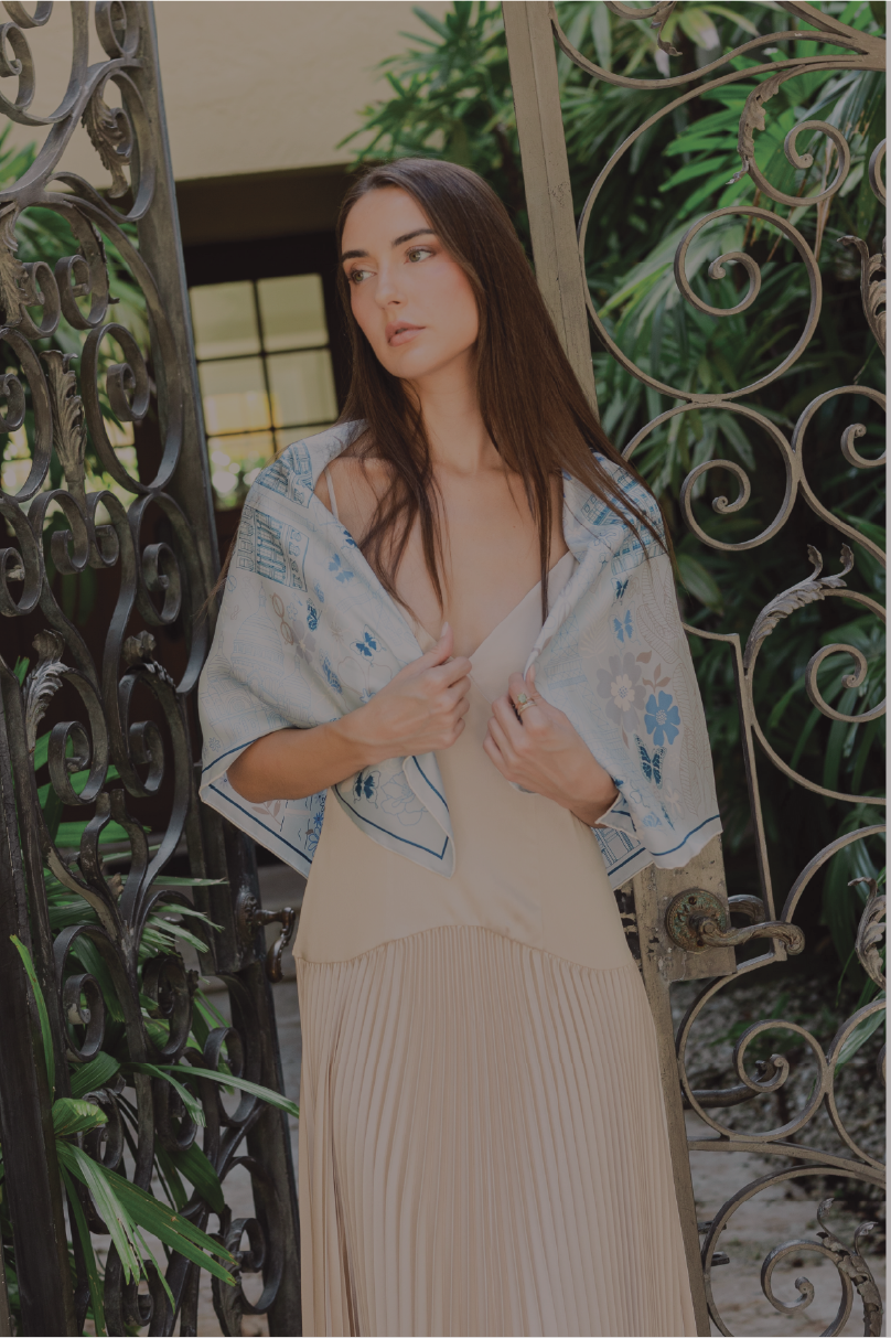 A woman wearing a beige pleated maxi dress and a light blue paris inspired silk scarf draped over her shoulders, standing in front of an ornate black wrought iron gate with green foliage in the background.