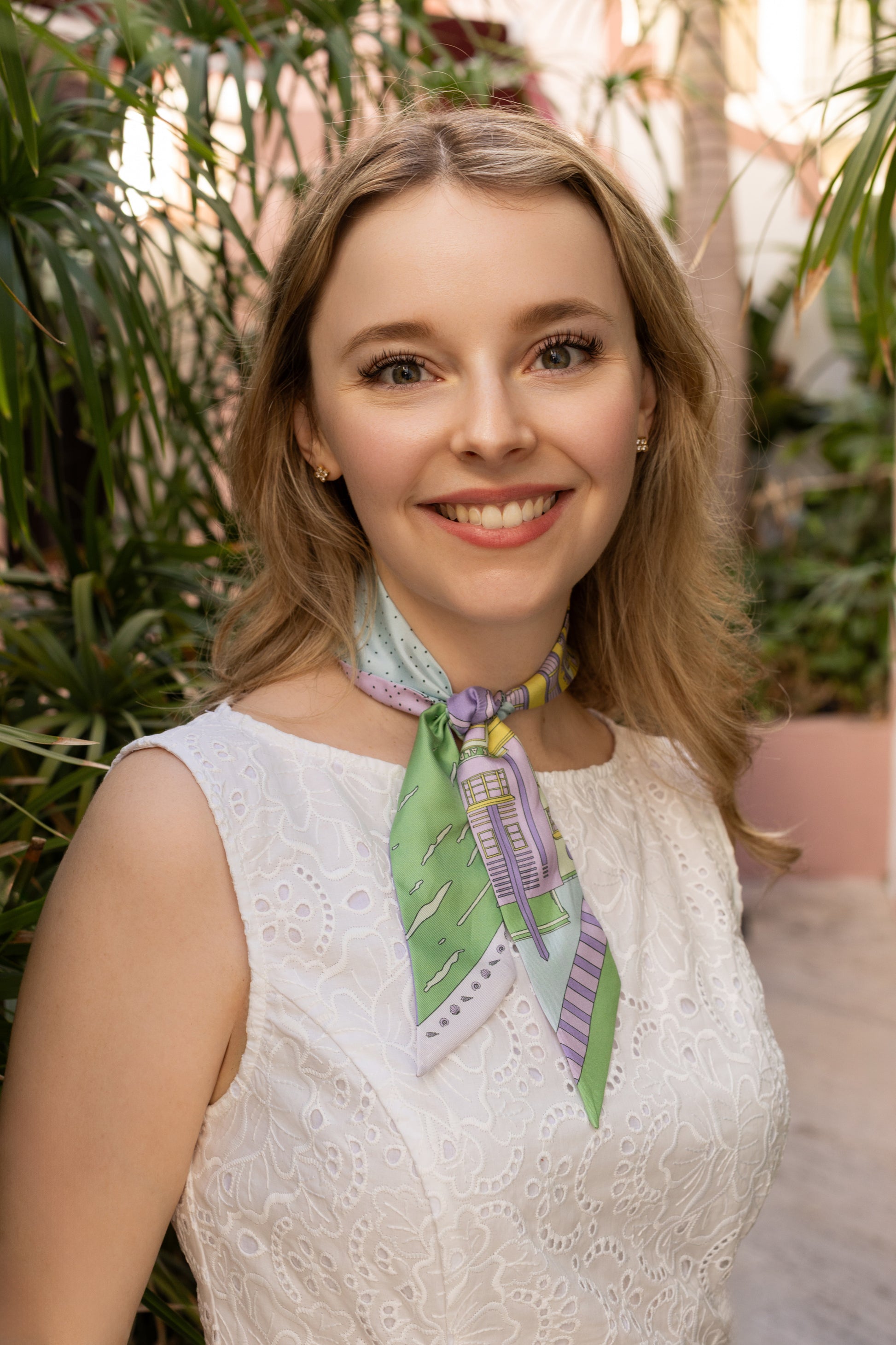 Woman wearing the Pastel Miami Beach silk twilly as a neck scarf with a white dress, featuring hand-drawn Miami Beach Art Deco architecture and coastal-inspired artwork in pastel purple, yellow, blue, and green tones.