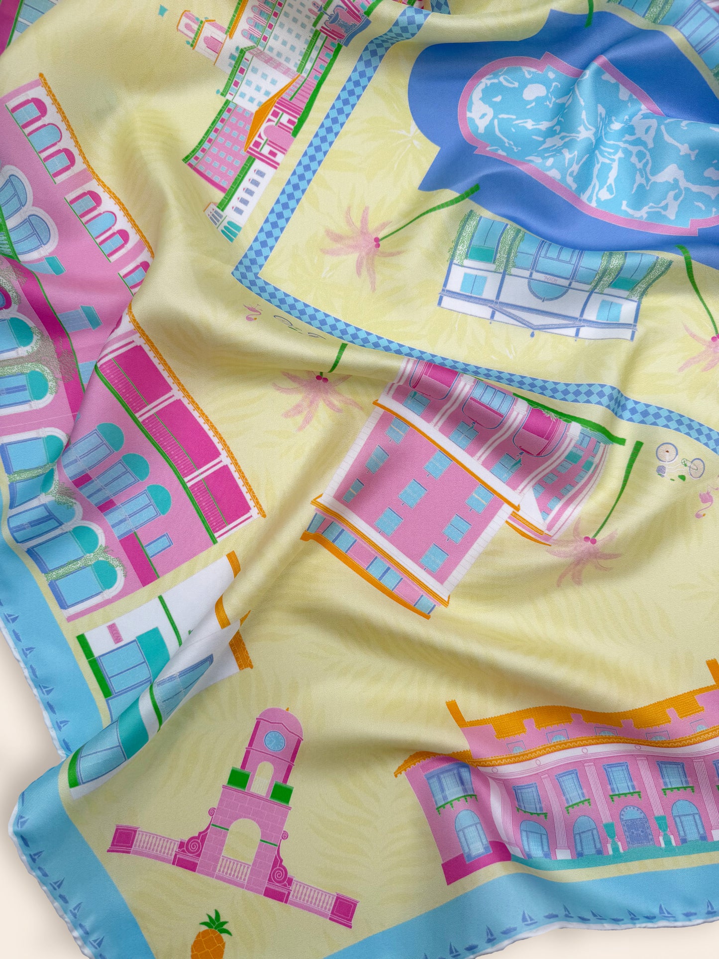 Close-up of the Pink Breeze Palm Beach silk scarf showcasing hand-drawn illustrations of The Breakers, The Colony Hotel, Worth Avenue, Royal Poinciana Plaza, and other iconic Palm Beach landmarks on a bright yellow background with pink, blue, orange and white details.
