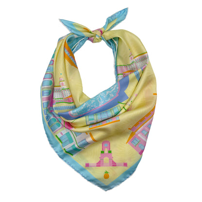 Pink Breeze Palm Beach silk scarf styled as a neck bandana featuring hand-drawn Palm Beach landmarks, architecture, and coastal-inspired artwork in bright yellow, pink, and blue tones.