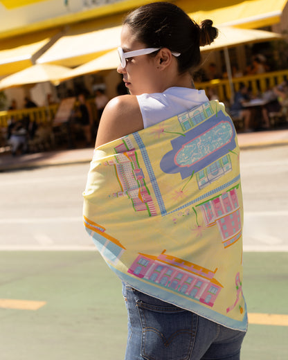 Woman wearing the Pink Breeze Palm Beach silk scarf as a shawl featuring hand-drawn Palm Beach landmarks including The Breakers, Worth Avenue, and the Palm Beach Clock Tower in bright yellow, pink, and blue tones.