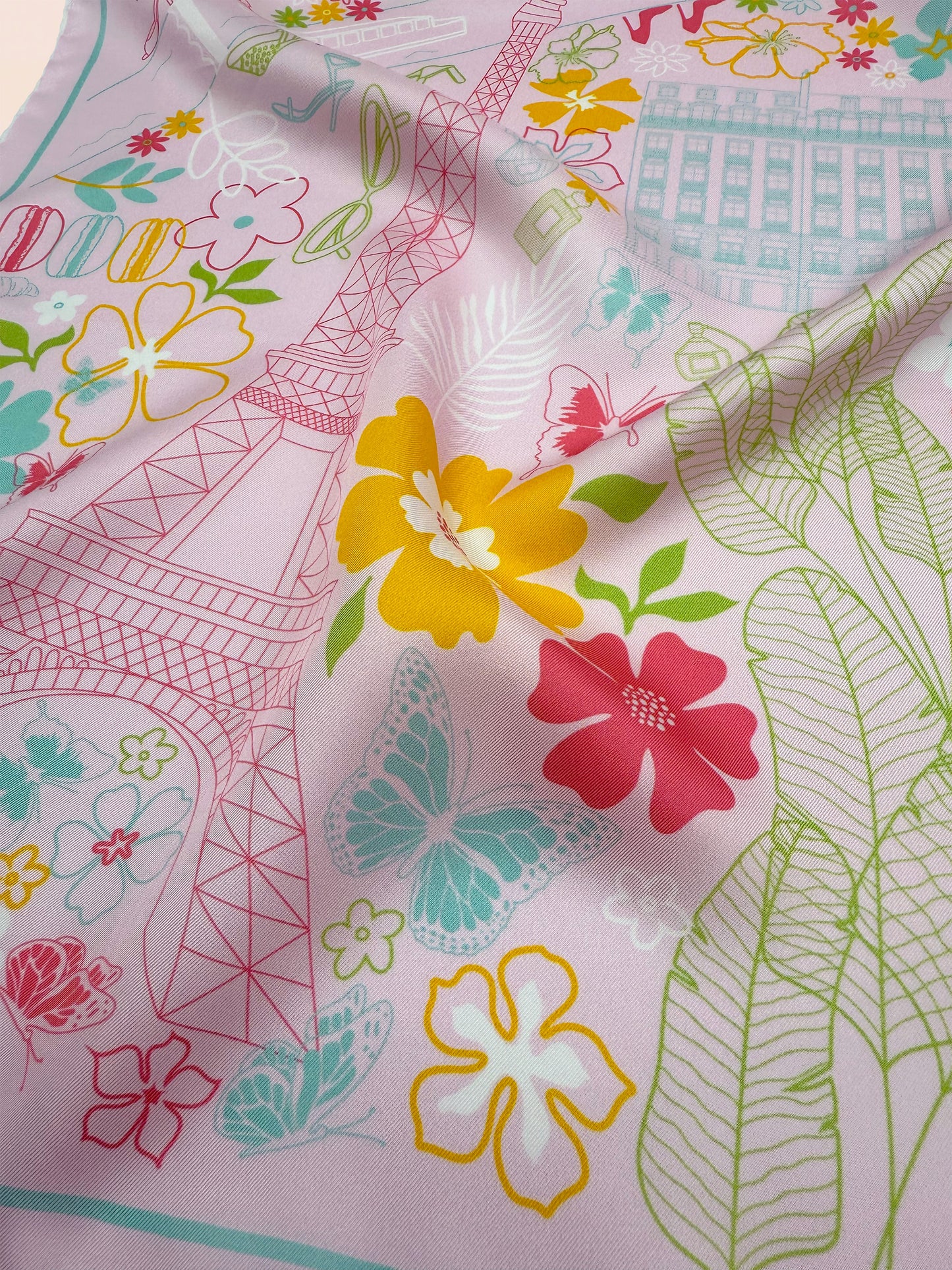 Close-up of the Rose Allure Paris silk scarf showcasing a hand-drawn map of Paris, iconic landmarks, fashion-inspired details, and colorful blue, green, and yellow accents on a light pink background.