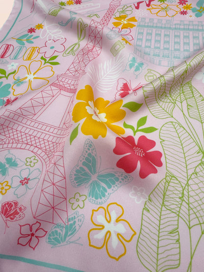 Close-up of the Rose Allure Paris silk scarf showcasing a hand-drawn map of Paris, iconic landmarks, fashion-inspired details, and colorful blue, green, and yellow accents on a light pink background.