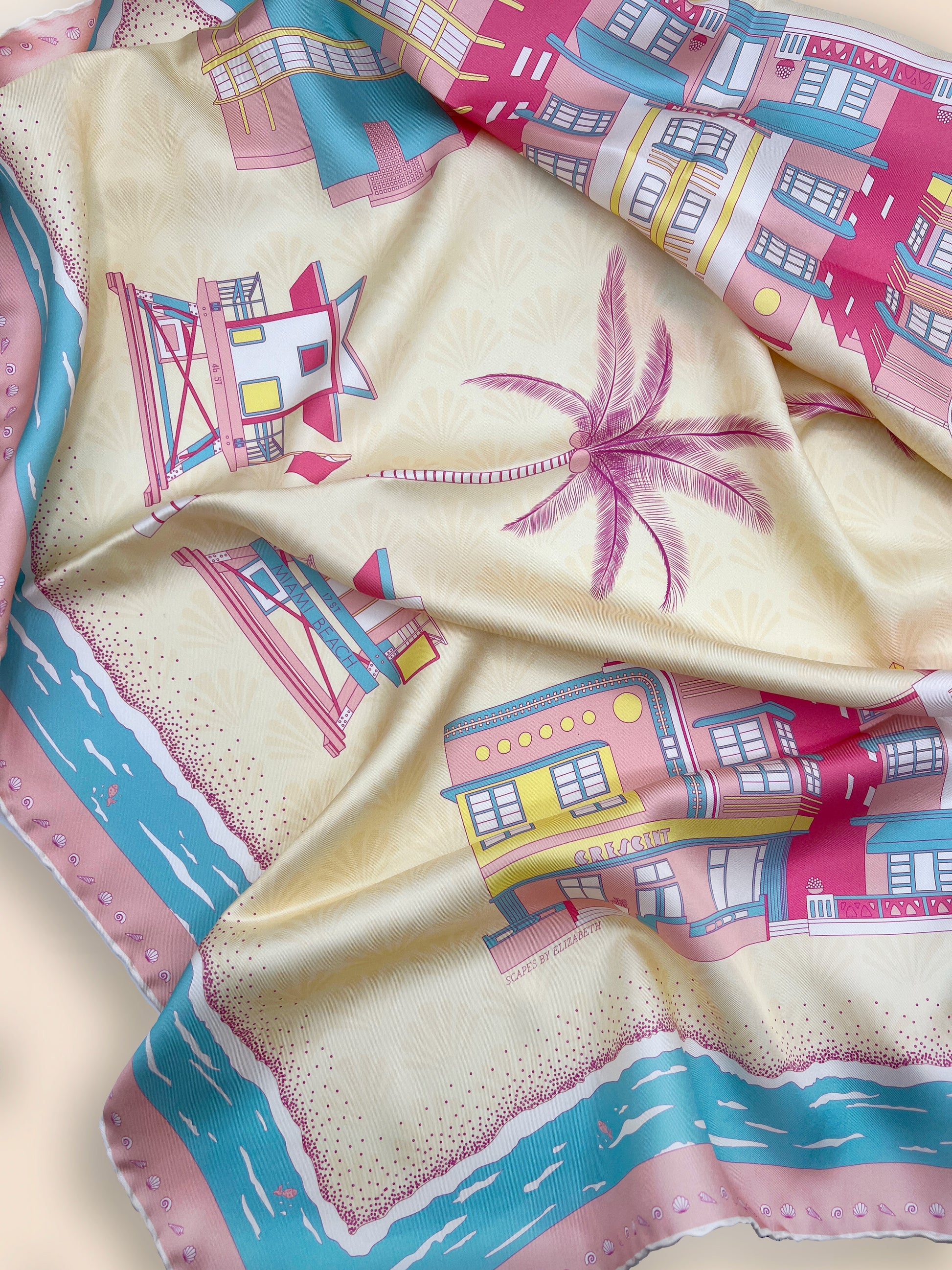 Close-up of the Rosy Splash Miami Beach silk scarf showcasing hand-drawn Art Deco architecture, iconic miami beach lifeguard towers, palm trees, a subtle shell pattern, and a decorative seashell border.