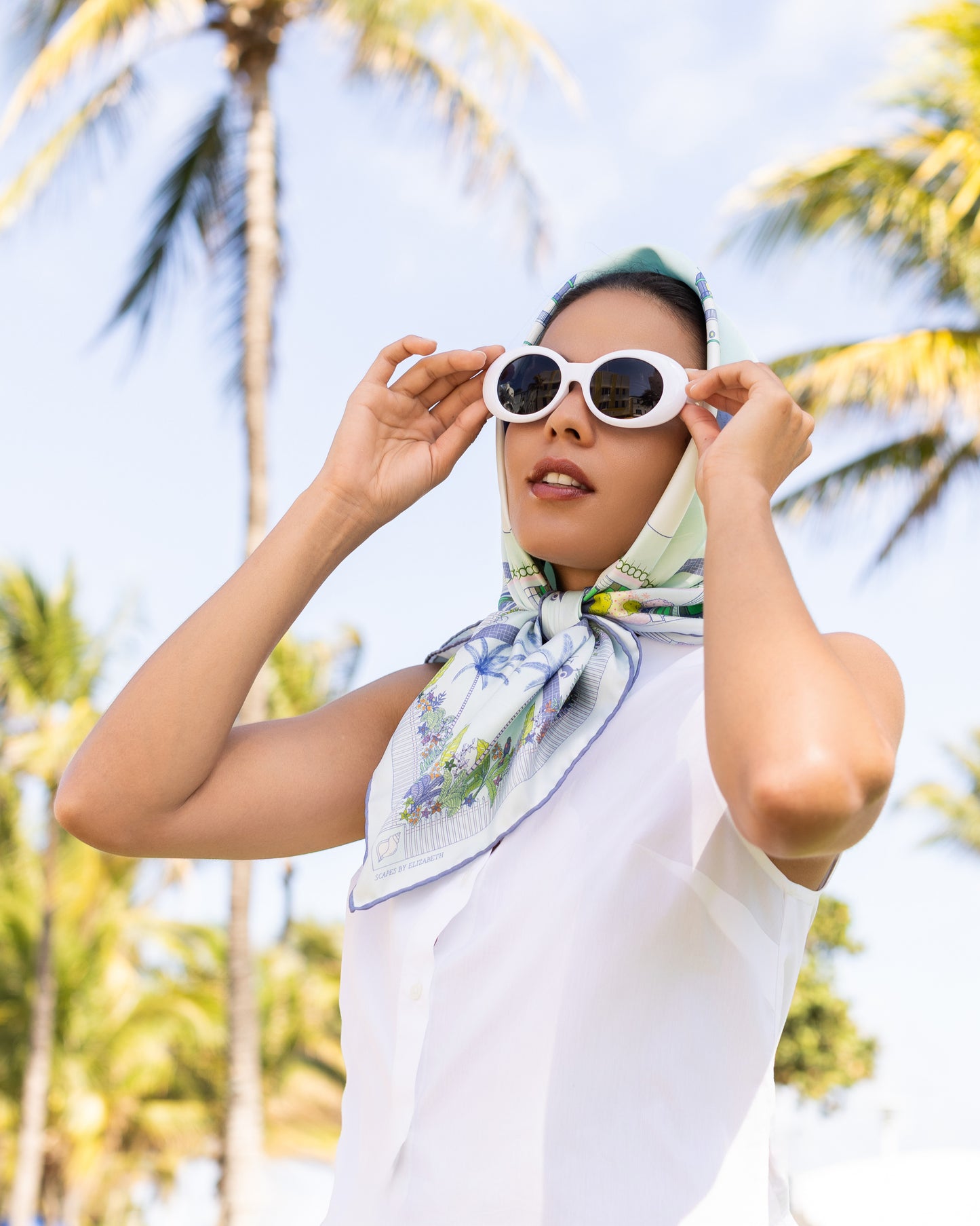 Woman wearing the Seafoam Blue Key West silk scarf as a head scarf featuring hand-drawn Key West architecture, Duval Street landmarks, tropical florals, and coastal-inspired artwork in shades of blue and green.