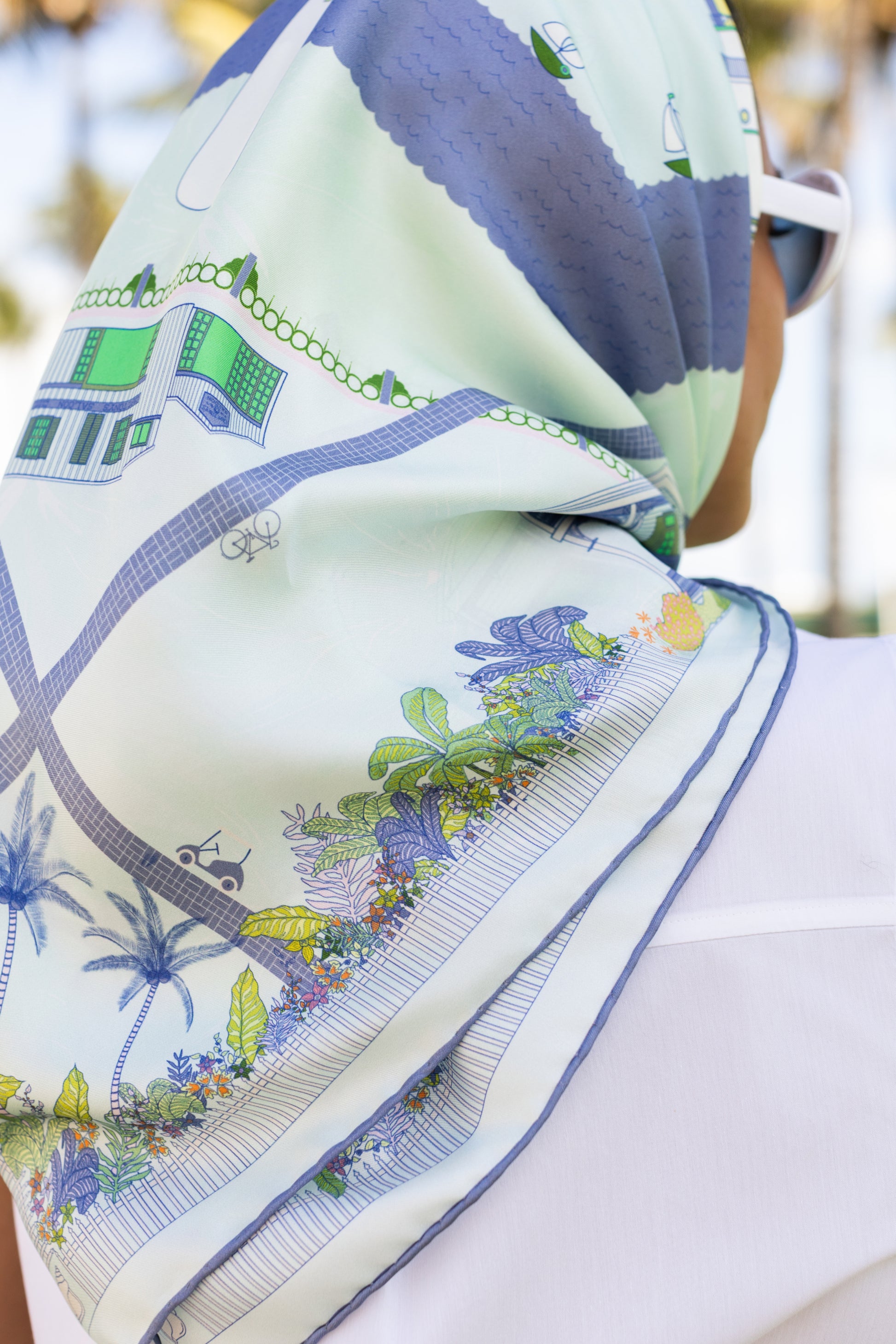 Close-up of the Seafoam Blue Key West silk scarf showcasing hand-drawn tropical flowers, lush landscaping, palm trees, and coastal-inspired artwork in shades of blue and green.