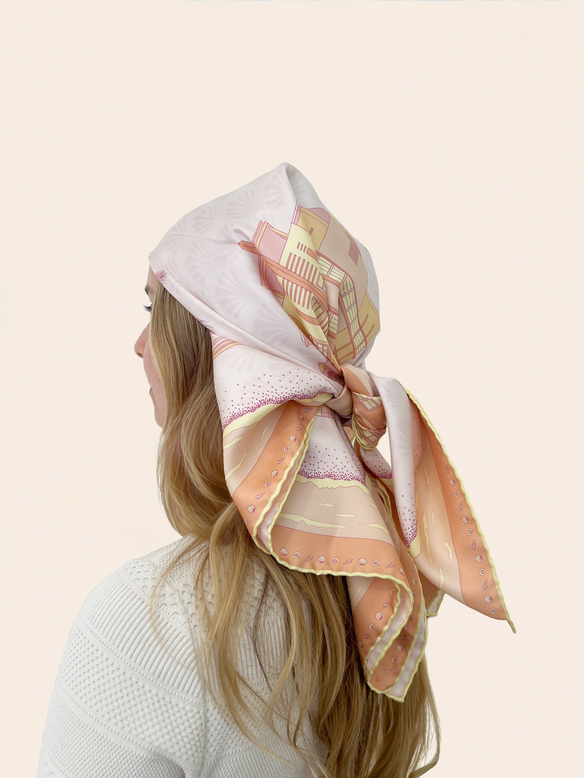 Woman wearing the Sunset Miami Beach silk scarf as a head bandana featuring hand-drawn miami beach Art Deco buildings, miami beach lifeguard towers, palm trees, and coastal scenery in sunset-inspired colors of pink, yellow, and orange.